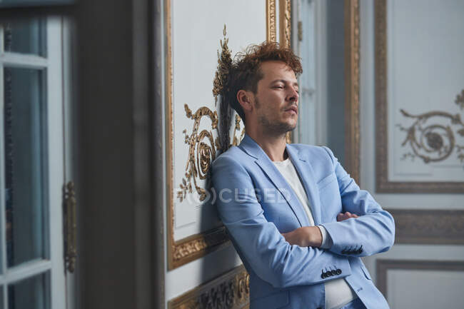 Pensativo hombre de traje de pie con los brazos cruzados y apoyado en la pared en la habitación elegante - foto de stock
