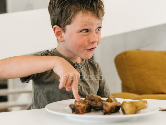 Позитивный ребенок сидит за столом с тарелкой с мясом и вкусный обед на кухне дома — стоковое фото
