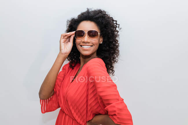 Trendy giovane afroamericana femminile con i capelli ricci in rosso usura e occhiali da sole guardando la fotocamera — Foto stock