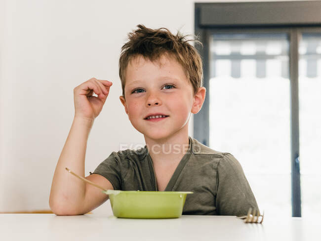 Retrato de adorable niño alegre sentado a la mesa a la hora del almuerzo en casa - foto de stock