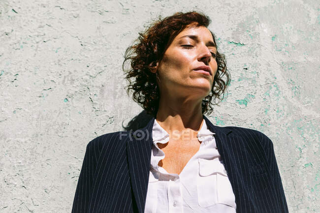 Femme réfléchie portant des vêtements élégants debout la lumière du soleil sur un mur minable avec les yeux fermés — Photo de stock