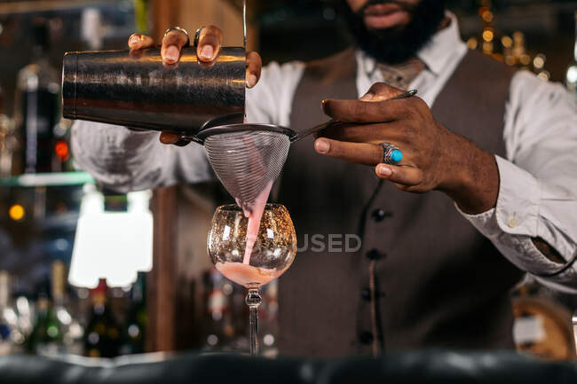Crop anonymous trendy ethnic male barkeeper pouring beverage from shaker into sieve above glass while preparing cocktail in bar — Stock Photo
