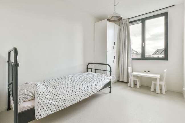 Einfaches minimalistisches Interieur mit weißem Schlafzimmer mit Einzelbett und Kleiderschrank in der Nähe des Fensters und kleinem Tisch mit Stühlen in einer modernen Wohnung — Stockfoto
