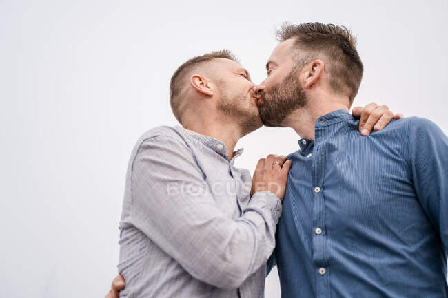 Happy man with modern haircut laughing while kissing with homosexual partner in shirt in daytime — Stock Photo