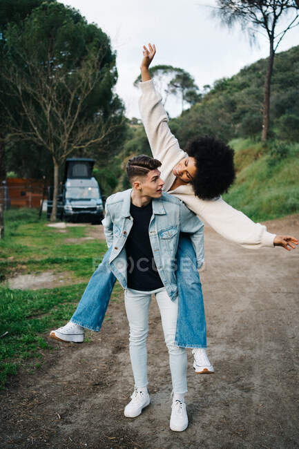 Young man giving cheerful ethnic girlfriend piggyback ride while looking away against camper and Thai Ridgeback on pathway — Stock Photo