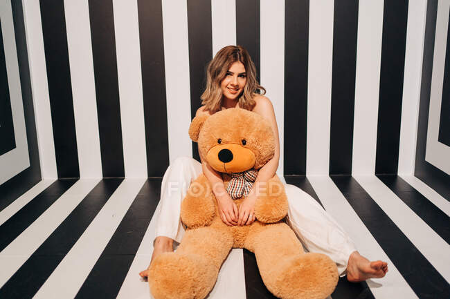 Cheerful barefoot woman in white pants with soft bear sitting on striped floor while looking at camera — Stock Photo