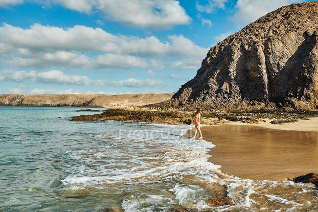 Turista femenina parada cerca de olas marinas espumosas en la playa de arena húmeda contra acantilados rocosos y cielo azul nublado durante las vacaciones de verano en Fuerteventura, España - foto de stock