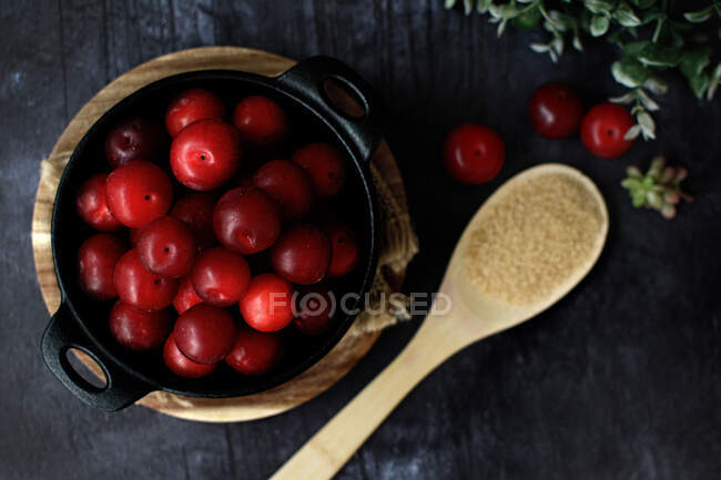Top view of bowl with fresh sweet plums served on black table with wooden spoon with sugar — Stock Photo