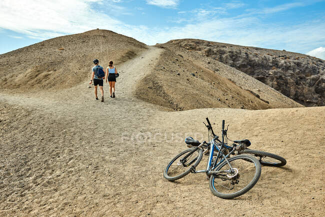 Hombres y mujeres irreconocibles caminando lejos de las bicicletas en el sendero arenoso que conduce a la colina seca en el día de fin de semana en Fuerteventura, España - foto de stock