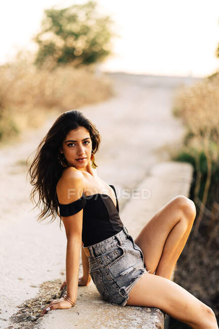 Side view of carefree barefoot young Hispanic female in black top and gray denim shorts sitting on stone bridge in summer day in countryside — Stock Photo