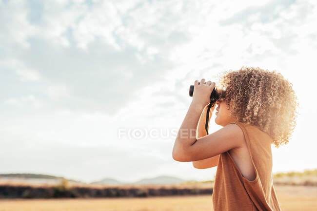 Side view of ethnic child with Afro hairstyle looking through binoculars and celebrating victory with raised arms while standing in dried filed in summer on sunny day and having fun — Stock Photo