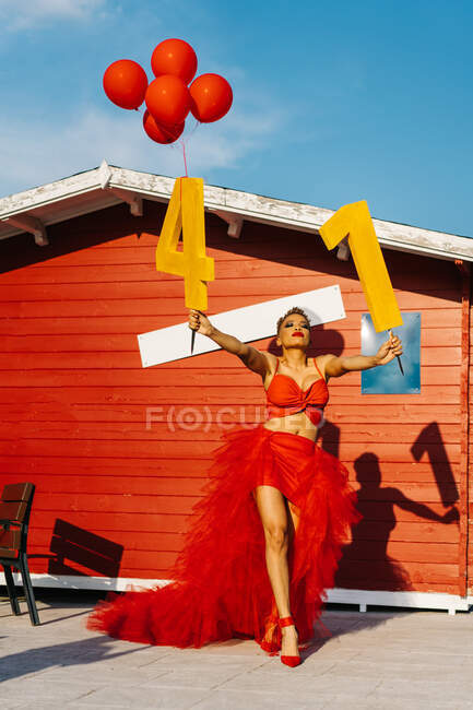 Модная черная женщина с декоративными номерами и красными воздушными шарами, стоящими на дорожке против строительства во время празднования дня рождения в солнечном свете — стоковое фото