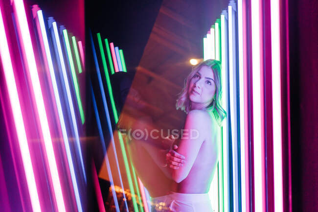 Side view of young dreamy woman reflecting between rows of glowing neon tubes while looking up — Stock Photo