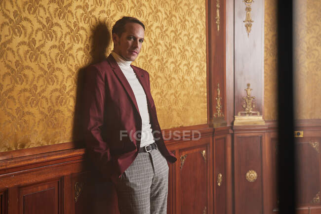 Уверенный взрослый актер в элегантной стильной одежде, держа руку в кармане и вдумчиво глядя в сторону, стоя у стены в винтажном стиле комнате — стоковое фото
