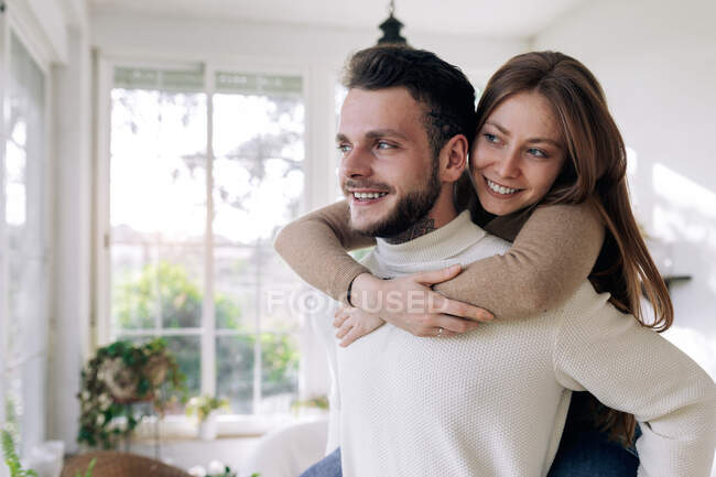 Smiling woman riding piggyback on boyfriend while against windows at home — Stock Photo
