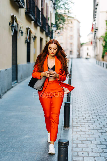 Stylish female with red hair and in vibrant red suit text messaging on smartphone in city street — Stock Photo