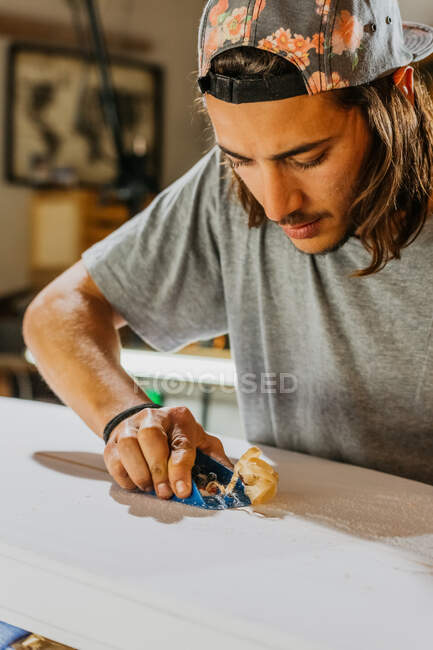 Master maschio focalizzato utilizzando il piano della mano e modellando la superficie liscia della tavola da surf in officina — Foto stock