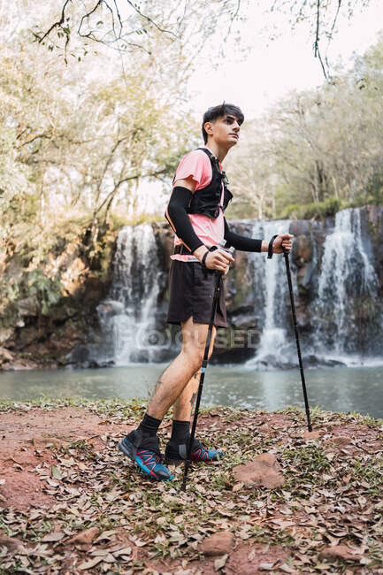 Seitenansicht eines männlichen Reisenden mit Trekkingstöcken, der in der Nähe eines Sees mit Wasserfall im Wald steht — Stockfoto