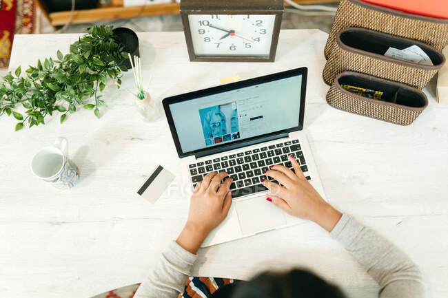 De cima de feminino usando netbook durante compras on-line enquanto sentado à mesa com cartão de plástico em casa e escolhendo bens — Fotografia de Stock