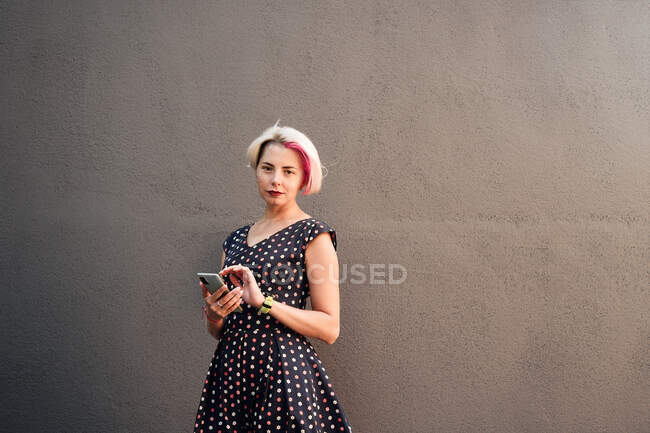 Delighted informal female with short hair and in dress messaging on mobile phone while standing against gray wall in street and looking at camera — Stock Photo