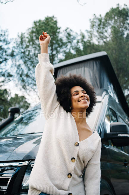 Alegre joven viajera afroamericana con ropa casual riendo felizmente mientras se apoya de nuevo en la caravana estacionada en la naturaleza durante las vacaciones de verano - foto de stock