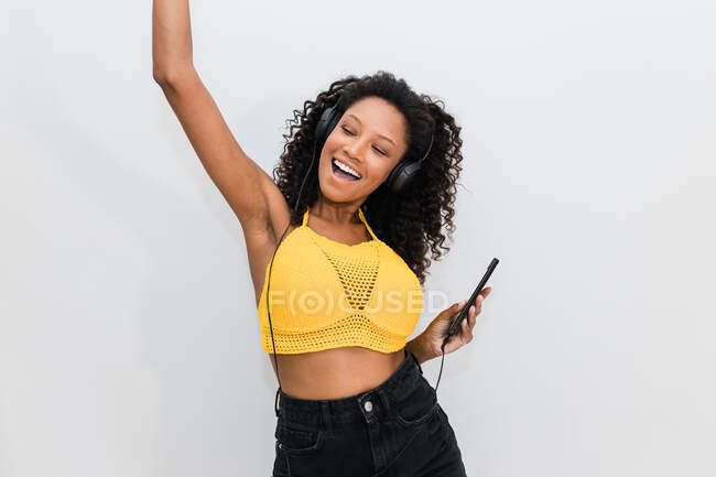Felice femmina afroamericana in cuffia con il cellulare che balla ascoltando la canzone su sfondo chiaro — Foto stock