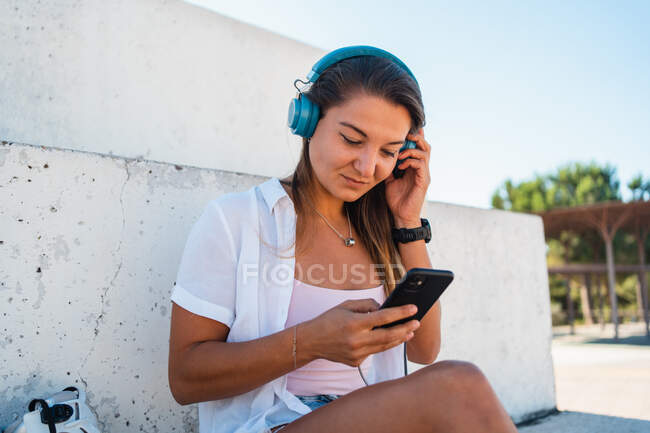 Positive cheerful young female browsing on mobile phone listening music on headphones on sunny day in summer in city — Stock Photo