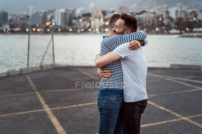 Sincere man embracing unrecognizable homosexual partner while looking away against lake and mount in town — Stock Photo
