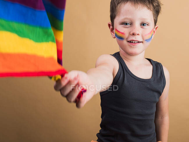Enfant joyeux avec maquillage sur les joues avec drapeau LGBTQ tout en regardant la caméra sur fond beige — Photo de stock