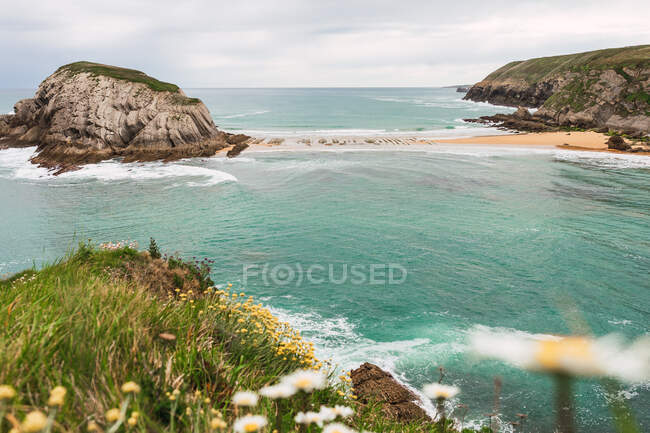 Paisaje fascinante con pequeña península rocosa y playa de arena bañada por agua de mar turquesa espumosa en Liencres Cantabria España - foto de stock