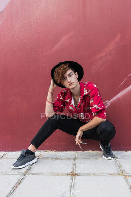 Homem homossexual na moda com unhas compridas em camisa ornamental olhando para a câmera enquanto se agacha no pavimento contra a parede — Fotografia de Stock