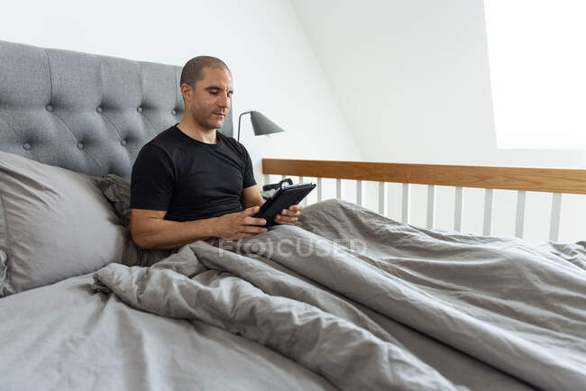 Calme mâle assis sur le lit sous la couverture et en utilisant la tablette après le réveil dans la chambre le matin à la maison — Photo de stock