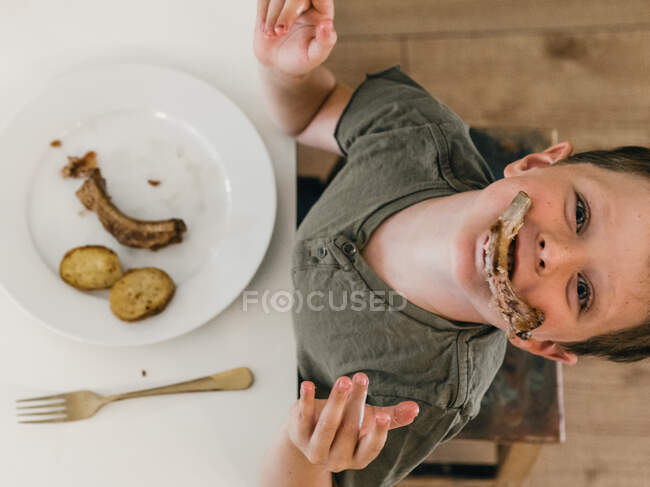 Top view of playful child with pork rib in mouth having lunch at table in kitchen and looking at camera — Stock Photo
