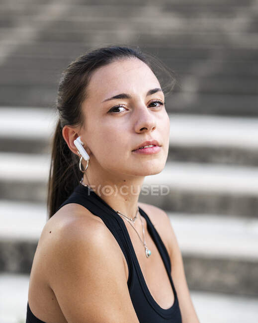 Jovem atleta feminina de sportswear preto ouvindo música em verdadeiros fones de ouvido sem fio e olhando para a câmera na rua da cidade — Fotografia de Stock