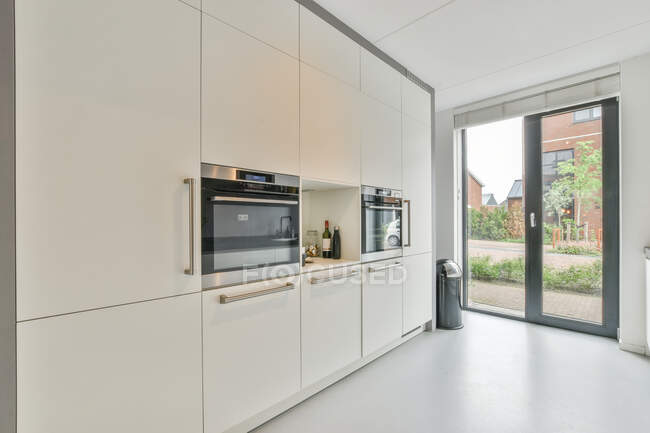 Минималистский стиль интерьера современной кухни с белыми встроенными шкафами и электроприборами возле стеклянной двери в доме — стоковое фото