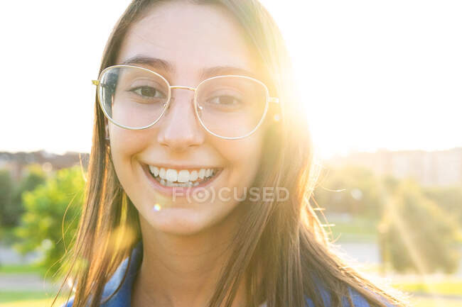 Positive young female in trendy clothes standing on green hill on background of city on sunny day and looking at camera in back lit — Stock Photo