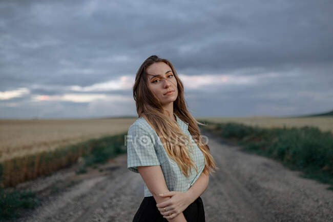 Vista lateral de la joven hembra consciente mirando la cámara en el camino cerca del prado bajo el cielo nublado en la noche en el campo - foto de stock