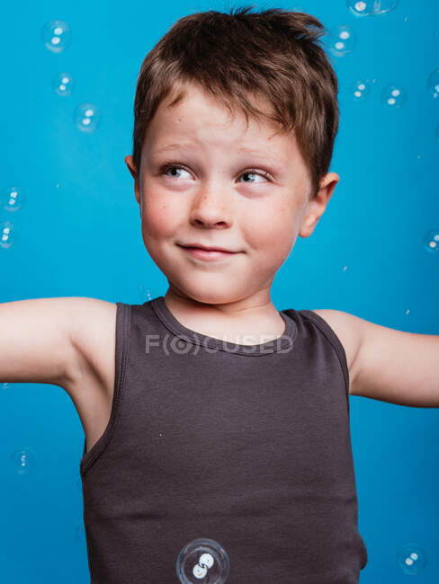 П'ятнадцятирічний хлопчик дивиться в студію з літаючими мильними бульбашками на синьому фоні — стокове фото