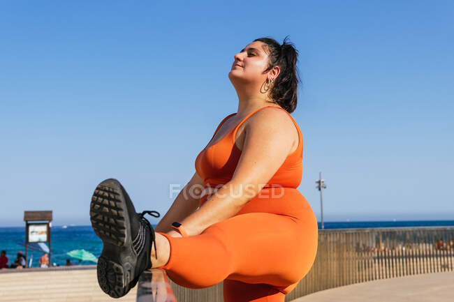 Vista lateral de atleta femenina étnica con cuerpo curvilíneo ejercitando mientras ojos cerrados en ciudad soleada - foto de stock