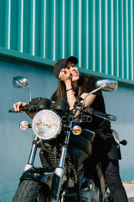 Motociclista positiva sentada en moto estacionada en la calle de la ciudad en un día soleado y mirando a la cámara - foto de stock