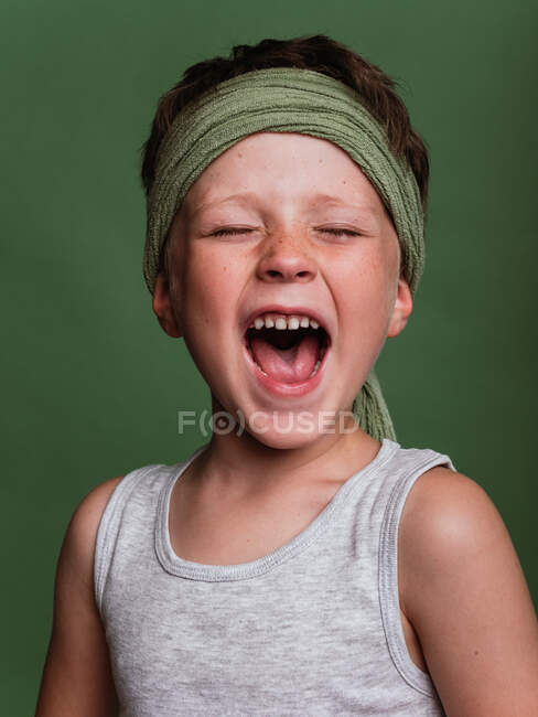 Positive preteen karate boy in hachimaki headscarf loudly shouting with closed eyes in studio on green background — Stock Photo