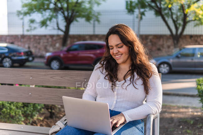 Fröhliche junge lateinamerikanische Studentin blättert am Laptop, während sie sich an einem Sommertag auf einer Holzbank in der Stadtstraße ausruht — Stockfoto