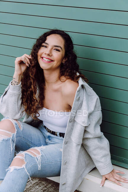 Charming female in stylish wear sitting near wall of building and leaning on hand while smiling and looking at camera — Stock Photo