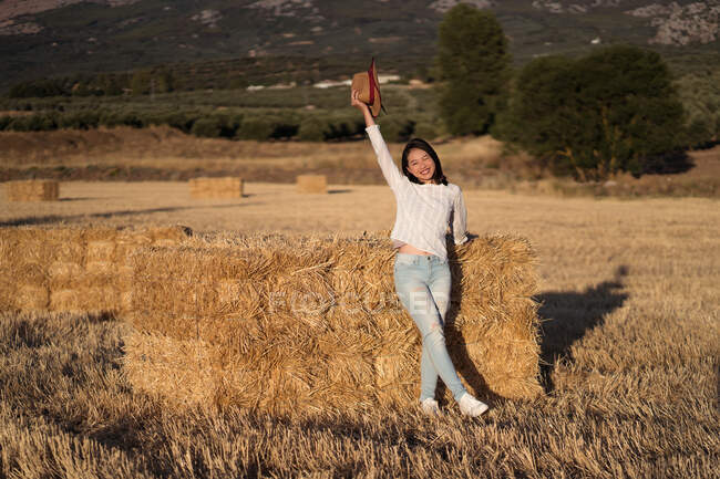 Содержание этнической женщины с соломенной шляпой, стоящей возле стога сена в поле и смотрящей в камеру — стоковое фото