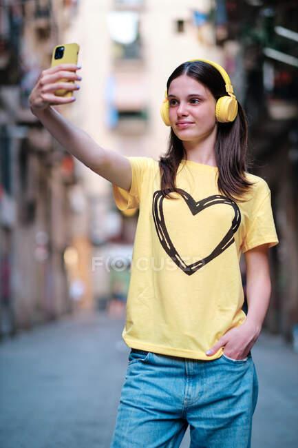 Carefree female in yellow headphones listening to music and taking self portrait on smartphone having fun in city street in summer — Stock Photo