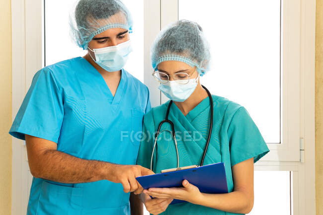 Серьезные врачи мужского и женского пола в масках читают отчет на планшете, стоя в коридоре клиники — стоковое фото