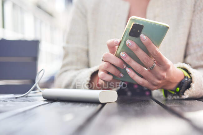 Анонімні альтернативні жінки з коротким волоссям, які переглядають соціальні медіа на смартфоні, сидячи за столом у вуличному кафе в сонячний день — стокове фото