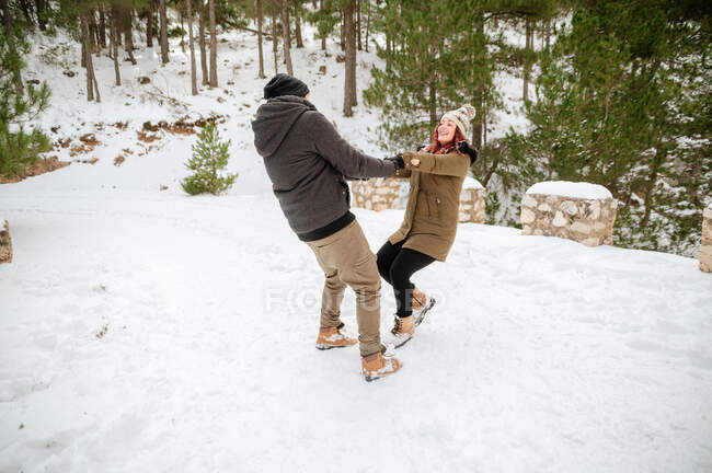Задоволена пара в теплому одязі, тримаючись за руки і кружляючи навколо в засніжених зимових лісах, розважаючись — стокове фото