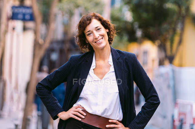 Позитивная женщина в стильной одежде идет по улице улыбаясь, глядя в камеру — стоковое фото