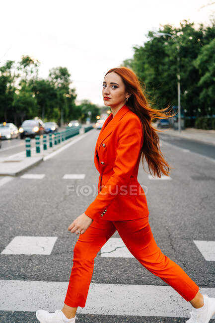 Vista lateral da mulher confiante com cabelo longo gengibre e na moda vívida laranja terno cruzando estrada na cidade e olhando para a câmera — Fotografia de Stock
